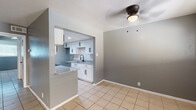 This single-floor space offers a practical and modern layout finished with light beige ceramic tile flooring throughout. The open living and dining area is spacious and bright, featuring large windows with horizontal blinds that allow ample natural light to fill the room. A ceiling fan with an integrated light fixture is centrally positioned overhead, and the walls are painted in a cool, medium gray tone that carries consistently throughout the unit.

The kitchen is a standout feature, designed in a galley-style configuration with white shaker cabinetry accented by matte black hardware. Granite countertops with a speckled gray finish extend along both sides of the kitchen and continue onto a peninsula that opens toward the dining area, creating a convenient pass-through. A hexagonal white tile backsplash adds visual texture, while stainless steel appliances — including a range and microwave — complete the functional layout. Recessed lighting illuminates the kitchen workspace effectively.

The bathroom includes a granite-topped vanity with a white shaker base cabinet, a wall-mounted mirror, and tile flooring consistent with the rest of the unit. The overall aesthetic is clean, cohesive, and move-in ready.
