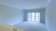 This single-floor residence features a spacious, open living area finished with soft, neutral-toned wall-to-wall carpeting that lends warmth underfoot. The room is defined by elegant crown molding that runs along the ceiling perimeter, adding a refined architectural detail to the otherwise clean, understated design. Light gray walls create a calm, neutral backdrop well-suited to a variety of interior styles.

A prominent set of sliding glass doors serves as the focal point of the main living space, flooding the interior with natural light and providing direct access to a private balcony enclosed by a metal railing. The balcony outlook offers views of the surrounding building exterior, giving the space a sense of connectivity to its environment. Sheer white curtains frame the doors, softening the natural light that enters throughout the day.

Recessed lighting in the ceiling supplements the abundant natural light, ensuring the space remains well-lit during evening hours. White baseboards provide a crisp contrast to the carpeted floors, contributing to the overall clean and polished aesthetic. The open layout allows for flexible furniture arrangements, making the living area highly functional and adaptable.