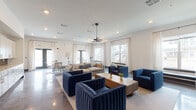 Located at 2303 Lebanon Pike in Nashville, Tennessee, this single-story clubhouse offers a well-designed communal space that balances modern aesthetics with functional comfort. The open-concept layout is anchored by polished concrete floors that reflect natural light streaming in through multiple large windows dressed in sheer white curtains, creating a bright and welcoming atmosphere throughout.

The main lounge area is generously proportioned, furnished with richly upholstered navy blue channel-tufted sofas and accent chairs arranged around a solid wood coffee table. A statement pendant light fixture with a geometric linear design hangs overhead, complementing recessed lighting throughout the ceiling. One wall features a textured chevron-patterned tile accent wall with a mounted flat-screen television, adding a striking focal point to the space.

A separate casual seating zone includes woven cantilever chairs and an additional lounge area, offering flexible configurations for residents to gather. French doors provide direct access to the exterior, enhancing connectivity to the outdoors.

The clubhouse also includes a dedicated kitchenette, outfitted with light wood cabinetry, open shelving, a sink, and a metallic three-dimensional tile backsplash that adds visual interest. The overall design presents a cohesive, contemporary aesthetic suited for community engagement.