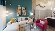 This single-floor residence makes a striking first impression with its dramatic high ceilings and bold design choices. The living area is defined by a deep teal accent wall adorned with vivid, large-scale portrait artwork, creating a gallery-like atmosphere. A neutral sectional sofa, a fuchsia accent chair, and a glass coffee table complete the space, which opens through floor-to-ceiling sliding glass doors to a private outdoor balcony with exterior views. Teal drape panels frame the generous natural light that floods the room.

The kitchen is a standout, featuring dark espresso cabinetry extending to impressive heights, crisp white countertops, wood-look plank flooring, and a full suite of stainless steel appliances. A large island with bar seating provides both workspace and a casual dining option, illuminated by pendant lighting beneath soaring ceilings.

The primary bedroom is an elegant retreat styled with a mirrored geometric headboard, deep purple accent tones, and multiple windows fitted with blinds and drapes. The en-suite bathroom offers a glass-enclosed shower and access to a walk-in closet. A second bedroom features its own teal accent wall, ample natural light, and plush carpeting. Its accompanying bathroom includes a soaking tub and a dedicated closet space with built-in shelving.