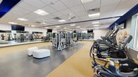 This fitness center offers a generously sized, single-floor layout designed to accommodate a full range of workout activities. The open floor plan is thoughtfully organized into distinct training zones, each featuring durable, sport-specific flooring — a combination of resilient rubber matting and rubberized cork surfaces — that provides both safety and comfort underfoot.

The cardio zone is lined with a row of modern treadmills and elliptical machines positioned along a wall adorned with a bold geometric triangle mural in orange and white, complemented by large windows that bring in natural light and outdoor views. Wall-mounted flat-screen TVs are positioned throughout for entertainment during workouts.

The strength and functional training areas house a comprehensive selection of selectorized cable machines, multi-station cable rigs, free weight racks, benches, and resistance training equipment. Full-length mirrors line multiple walls, enhancing the sense of openness and allowing users to monitor their form. Ceiling fans are distributed evenly throughout to maintain airflow, supplemented by a recessed drop-ceiling grid with bright panel lighting.

A welcoming entry area features stylish white cushioned ottomans, open wooden shelving units for personal storage, and a wall-mounted water station. The overall aesthetic is clean and modern, with a neutral gray and white palette accented by navy blue architectural details.