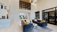 The Fitzgerald's lobby makes a striking first impression through its dramatic double-height ceilings and expansive floor-to-ceiling steel-framed windows that flood the space with natural light. Polished large-format marble tile flooring extends throughout, lending an elegant, refined quality to the entire area. The main seating area anchors the lobby with deep navy velvet sofas accented by bold yellow throw pillows, arranged around round marble-topped coffee tables atop a textured area rug — creating a welcoming and stylish atmosphere. A built-in workstation niche along one wall features a dark wood and stone counter, paired with wood barstools and dual monitors, offering a functional resource for residents. Adjacent to the main lounge, a more intimate seating nook accommodates a wall-mounted flat-screen television, a warm walnut credenza, and a pair of upholstered lounge chairs — ideal for relaxed viewing. The reception desk, finished in white marble, sits prominently near the entrance, while a steel-framed glass door leads to additional spaces beyond. Large planters with lush greenery frame the main entry doors, softening the modern aesthetic. A contemporary chandelier completes the grand, airy ambiance of this well-appointed lobby.