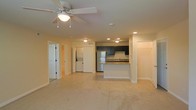This single-floor residential unit offers a thoughtfully designed open-concept layout that seamlessly connects the living area to the kitchen, creating a spacious and functional environment. The living area features soft carpeted flooring and is fitted with a ceiling fan and light fixture, providing both comfort and practicality. Recessed lighting throughout enhances the warm, neutral ambiance of the space.

The kitchen is a standout feature, showcasing rich dark cabinetry that contrasts elegantly with light countertops. A peninsula-style breakfast bar adds both prep space and a casual dining option. Stainless steel appliances, including a refrigerator and dishwasher, complete the kitchen's modern and functional appeal. A sink with a window-adjacent position adds to the kitchen's bright and airy feel.

A dedicated laundry area is conveniently located just off the main living space, housing a stacked washer and dryer unit behind a full-size door, keeping household tasks neatly tucked away. Additional doorways lead to private rooms and hallways, suggesting well-separated living and sleeping quarters. The clean, neutral color palette, recessed lighting, and open sightlines throughout give the unit a welcoming and move-in-ready character.