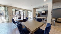 This single-floor community clubroom is thoughtfully designed to offer a variety of social and recreational experiences within one open, well-appointed space. The layout is divided into distinct functional zones that flow seamlessly into one another, unified by light wood-look tile flooring and a cohesive contemporary design palette of navy blue, warm neutrals, and slate gray accents.

The dining and collaboration area features multiple large rectangular tables paired with tufted navy blue accent chairs with chrome legs, creating an upscale yet inviting atmosphere. Large floor-to-ceiling windows and glass-panel doors flood the space with natural light and offer views of the surrounding outdoor areas, complemented by patterned curtains that add a pop of color and texture.

The lounge and gaming zone is anchored by a plush navy sectional sofa, bold geometric pendant lighting in brushed gold, and a wall-mounted flat-screen TV. A full-size billiards table positioned near a window adds to the recreational appeal of this area.

A bar-height counter with seating along a textured accent wall serves as a snack or media station, complete with an additional mounted TV. Entry to the space is defined by sleek floor-to-ceiling glass-panel doors with dark metal frames, providing an elegant and transparent transition that hints at the vibrant interior within.