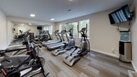 This fitness center offers a clean, modern environment designed to support a comprehensive workout experience. The space is finished with neutral-toned walls and striped, multi-toned carpet tiles that add subtle visual texture underfoot, transitioning to large-format tile flooring near the entryway corridor. Recessed and flush-mounted ceiling lights provide consistent, even illumination throughout.

The main workout area is outfitted with a lineup of cardio equipment, including Vision Fitness treadmills, elliptical cross-trainers, and stationary cycling bikes. A full-length wall mirror along one side visually expands the room while allowing users to monitor their form. A dumbbell rack with a weight bench supports strength training needs. A wall-mounted flat-screen television adds an entertainment option during workouts.

A row of large windows along one wall invites ample natural light and frames views of mature surrounding trees, creating a refreshing connection to the outdoors. The entryway corridor, finished in large tile, leads to a double-door entrance and additional adjoining rooms, suggesting space for changing or storage. The overall ambiance is functional yet polished, offering a well-organized and inviting environment for fitness.