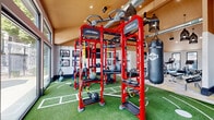 Located at 8131 E Colorado Ave in Denver, Colorado, this single-floor fitness center offers a thoughtfully designed exercise environment packed with professional-grade equipment. The space is defined by striking vaulted ceilings with warm wood paneling, exposed ductwork, and decorative pendant lighting that lend an inviting yet energetic atmosphere.

The main exercise area is divided into two distinct zones. A synthetic turf section, marked with field lines, anchors the space and hosts a bold red Life Fitness SYNRGY360 functional training rig, complete with TRX suspension straps, medicine balls, kettlebells, weight plates, and a TKO heavy punching bag — ideal for functional and high-intensity training. Large floor-to-ceiling windows on one side offer direct views of an outdoor pool area, flooding the turf zone with natural light.

The second zone features smooth, patterned tile flooring and houses a comprehensive selection of strength and cardio machines, including cable machines, a dumbbell rack with mirror wall, weight benches, treadmills, a Peloton spin bike, and an elliptical. Framed black-and-white photography adorns the walls, and a mounted flat-screen television adds convenience for members. Natural light streams in through multiple windows, creating a bright and motivating environment throughout.