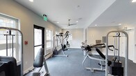 This fitness room offers a functional and well-organized space designed to accommodate a variety of workout routines. The room features clean white walls and light gray carpeted flooring, creating a neutral and focused environment conducive to exercise. Recessed lighting is complemented by a ceiling fan, ensuring the space remains well-lit and properly ventilated during use. Natural light filters in through multiple windows along one wall, adding brightness and an open feel to the interior.

The room is equipped with a multi-station cable machine, an adjustable weight bench, and what appears to be a dedicated pilates or stretching apparatus, offering users a range of strength and flexibility training options. The equipment is arranged to allow comfortable movement throughout the space. A mirrored or glass-paneled feature along one wall adds visual depth and reflects light, enhancing the overall sense of openness. An emergency exit sign and safety signage are visible, reflecting attention to building code compliance. Overall, the space presents a clean, modern, and practical fitness facility.