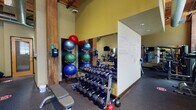Located at 1119 Mississippi Ave in St. Louis, Missouri, this fitness center blends historic architectural character with modern exercise amenities. The single-floor space is defined by exposed brick and stone walls, sturdy timber columns, and an open ceiling with visible wooden beams and ductwork — all contributing to a warm, industrial aesthetic. Natural light filters in through multiple large windows, brightening the workout environment.

The cardio zone is well-equipped with treadmills, elliptical trainers, and stationary bikes, complemented by wall-mounted televisions for entertainment during workouts. Full-length mirrors line sections of the walls, enhancing the sense of openness and allowing users to monitor their form. The strength training area features a comprehensive dumbbell rack with a wide range of weights, a stability ball storage rack, kettlebells, and a weight bench. A dedicated whiteboard serves as a space for workout planning and instructional notes.

A separate strength machine section houses cable and pulley systems alongside a multi-function weight machine, providing options for a full-body workout. Rubber flooring throughout the facility ensures safety and durability. The soaring ceiling height adds to the spacious, airy feel of the gym, making it a well-rounded and inviting space for fitness enthusiasts of all levels.