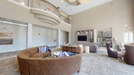 This elegant community clubhouse is defined by its dramatic double-height ceilings and an open, airy lounge that serves as the heart of the space. The main gathering area features a curved upper-level balcony with metal and wood railings that overlooks the seating below, adding architectural depth and grandeur. Multiple seating arrangements — including plush curved sofas, leather accent chairs, and glass-topped coffee tables — are thoughtfully arranged on patterned carpet tiles throughout the lounge, creating both communal and intimate settings. A built-in wood media wall with integrated shelving anchors one end of the room. Tall, floor-to-ceiling windows and sliding glass doors flood the interior with natural light and provide direct views of the outdoor pool and landscaped surroundings, seamlessly connecting the indoor and outdoor environments. A catering kitchen off the main lounge is outfitted with granite countertops, wood cabinetry, stainless steel appliances including a full-size refrigerator, open display shelving, and large windows that continue the bright, welcoming aesthetic. A carpeted staircase with sleek metal balusters and a warm wood handrail leads to the upper mezzanine level. Throughout, the neutral warm palette and contemporary furnishings create a polished, inviting atmosphere.