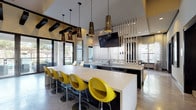 Located at 8405 Manderville Ln in Dallas, TX, this single-story community clubhouse delivers a sophisticated, contemporary aesthetic defined by a cohesive color palette of warm neutrals, bold yellows, and dark wood accents. The open social hub centers around a large kitchen island topped with a sleek white countertop and lined with vibrant yellow bar stools, all illuminated by a striking row of perforated pendant lights. A vertical metal slat partition elegantly separates the kitchen area from the adjoining space, adding architectural depth. The lounge area showcases a textured geometric feature wall framing a wall-mounted TV, complemented by a plush sectional sofa, decorative glass wall art, and a statement circular chandelier. Polished concrete floors run throughout the main gathering areas, lending an industrial-chic feel balanced by warm furnishings. A private media or sitting room offers a large window with patterned drapery, built-in dark cabinetry with a white countertop, and a mounted flat-screen TV — ideal for quieter gatherings. The fitness center is well-appointed with hardwood-style flooring, high ceilings, large mirrors, cardio equipment, free weights, a punching bag, and yoga space. Natural light flows in from multiple windows throughout, creating an open, welcoming atmosphere across the entire facility.