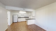 This single-story residence offers a practical and straightforward layout designed for comfortable everyday living. The open-concept kitchen and living area form the heart of the unit, where wood-look laminate flooring extends throughout. The kitchen is equipped with white cabinetry providing ample storage, dark speckled countertops, a double-basin sink, a white electric range, a dishwasher, and a full-size refrigerator. A peninsula with a matching dark countertop extends into the living area, creating a natural divide between the two spaces while offering additional prep or seating space. Overhead fluorescent lighting illuminates the kitchen efficiently. The living area is open and well-proportioned, with a textured ceiling throughout and a window fitted with vertical blinds allowing natural light to enter. The bedrooms feature neutral wall-to-wall carpet and windows with vertical blinds, keeping the rooms bright yet private. The bathroom includes a tiled shower-tub combination, a toilet, and wood-look tile flooring, maintaining a clean, functional aesthetic consistent with the rest of the unit. Throughout the space, the neutral white walls and light tones create a clean, move-in-ready environment.