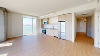Located at 39 Bruton Street in San Francisco, this modern 3-bedroom, 2-bathroom residence offers an open, light-filled layout across a single floor. The combined kitchen and living area serves as the heart of the home, featuring light wood-tone cabinetry, stainless steel appliances including a refrigerator, range, and microwave, a subway tile backsplash, and expansive floor-to-ceiling windows that frame breathtaking views of the San Francisco Bay and the Bay Bridge. Light hardwood flooring flows throughout, complementing the clean white walls and exposed concrete structural columns that lend an industrial-modern character to the space. The living area is generously proportioned, bathed in natural light and overlooking the bay. The primary bedroom is well-sized and features large windows with panoramic water views, including sightlines of the San Francisco skyline and Alcatraz Island. Additional bedrooms offer access to natural light and outdoor views. Two hallways connect the rooms efficiently, with two closets providing ample storage. The two bathrooms are finished in neutral tones, one featuring a floating wood vanity with a white countertop and a tiled tub-shower combination, the other with a classic tiled surround.