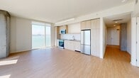 Located at 39 Bruton Street in San Francisco, this modern 3-bedroom, 2-bathroom residence offers an open, light-filled layout across a single floor. The combined kitchen and living area serves as the heart of the home, featuring light wood-tone cabinetry, stainless steel appliances including a refrigerator, range, and microwave, a subway tile backsplash, and expansive floor-to-ceiling windows that frame breathtaking views of the San Francisco Bay and the Bay Bridge. Light hardwood flooring flows throughout, complementing the clean white walls and exposed concrete structural columns that lend an industrial-modern character to the space. The living area is generously proportioned, bathed in natural light and overlooking the bay. The primary bedroom is well-sized and features large windows with panoramic water views, including sightlines of the San Francisco skyline and Alcatraz Island. Additional bedrooms offer access to natural light and outdoor views. Two hallways connect the rooms efficiently, with two closets providing ample storage. The two bathrooms are finished in neutral tones, one featuring a floating wood vanity with a white countertop and a tiled tub-shower combination, the other with a classic tiled surround.