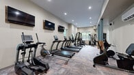Located at 1350 Gage St in Columbus, Ohio, this fitness center offers a thoughtfully designed workout space across a single floor. The layout is organized to accommodate a diverse range of exercise activities, making efficient use of the available floor area.

The cardio zone features a row of TRUE treadmills, an elliptical trainer, a rowing machine, and a stationary bike, all positioned along the main wall. Multiple flat-screen televisions are mounted at eye level, providing entertainment during workouts. Natural light filters in through several windows, complementing the bright recessed ceiling lighting that keeps the space well-illuminated throughout.

The strength training area includes a multi-functional cable machine, a power rack with an adjustable bench, and a dedicated free weights section. A large wall mirror enhances the sense of space and allows users to monitor their form. A medicine ball rack stocked with various weighted balls, resistance bands, and rolled yoga mats make the stretching and functional training zone a versatile area for floor exercises and mobility work.

The flooring consists of durable, patterned carpet tiles in warm earth tones, providing cushioning underfoot. Neutral beige walls and a clean, modern aesthetic give the space a polished, professional atmosphere. A water cooler and what appears to be a private room with frosted glass doors round out the amenities of this comprehensive fitness facility.