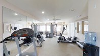 This fitness center offers a comprehensive workout environment designed to accommodate a range of exercise routines. The space is outfitted with an array of cardio equipment, including a treadmill, elliptical trainer, and stationary bike, alongside a multi-station cable machine for strength training. Large mirrors line one full wall, creating a sense of expanded space while allowing users to monitor their form during exercise. The light-colored flooring provides a clean, functional surface throughout the room. Natural light streams in through several windows dressed with decorative curtains, contributing to an open and inviting atmosphere. Recessed lighting and a centrally mounted ceiling fan ensure the space remains well-lit and properly ventilated during use. Built-in storage closets along one wall offer convenient organization for additional equipment or accessories. The neutral wall tones and bright ceiling give the room a clean, practical aesthetic. Overall, this fitness center is thoughtfully arranged to maximize usability while maintaining a comfortable and energizing environment for daily workouts.
