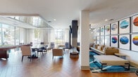 Located at 605 W Chapel Hill St in Durham, NC, this thoughtfully designed single-floor communal space blends modern aesthetics with comfortable functionality. The open-plan layout is anchored by striking herringbone-patterned wood flooring that flows seamlessly throughout, creating a warm and cohesive visual foundation.

At the heart of the space stands a dramatic double-sided fireplace with a dark surround, topped by a wall-mounted flat-screen television — serving as a natural divider between distinct seating zones. The lounge area features plush sofas and accent chairs arranged atop a vibrant teal area rug, with a curated gallery wall of bold, colorful circular artwork adding artistic character.

Floor-to-ceiling windows along one wall flood the space with generous natural light and provide open views of the surrounding streetscape, complemented by elegant sheer drapery. A dedicated dining area with a large table and upholstered seating takes advantage of this sun-drenched corner.

A separate kitchenette area is outfitted with dark wood cabinetry, a white quartz countertop island with teal bar stools, a marble-effect tile backsplash, and pendant lighting with gold accents — ideal for casual gatherings. Additional banquette seating and high-top tables offer flexible arrangements for work or socializing, making this a versatile and inviting communal environment.