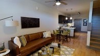 This well-appointed two-floor residence offers a thoughtfully designed layout that blends comfort and modern style. The main living area features warm hardwood flooring, neutral-toned walls, and a ceiling fan that adds both function and visual interest overhead. A plush leather sofa anchors the living space, complemented by a geometric-patterned area rug and a sleek glass coffee table with gold accents, creating an inviting and contemporary atmosphere.

The open-concept design flows seamlessly into a dining area, where a round bar-height table surrounded by green upholstered chairs serves as an elegant transitional space between living and kitchen. Pendant lights hang above, casting a warm glow across the area.

The kitchen is finished with dark espresso cabinetry, light countertops, and modern appliances, offering a rich contrast that adds depth to the overall aesthetic. A breakfast bar provides additional seating and further connects the kitchen to the social spaces of the home.

Throughout, the design emphasizes a cohesive color palette of warm browns, greens, and gold accents, lending the space a sophisticated yet comfortable character. Spanning two floors, this residence provides ample room for both relaxed living and functional everyday use.