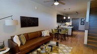 This well-appointed two-floor residence offers a thoughtfully designed layout that blends comfort and modern style. The main living area features warm hardwood flooring, neutral-toned walls, and a ceiling fan that adds both function and visual interest overhead. A plush leather sofa anchors the living space, complemented by a geometric-patterned area rug and a sleek glass coffee table with gold accents, creating an inviting and contemporary atmosphere.

The open-concept design flows seamlessly into a dining area, where a round bar-height table surrounded by green upholstered chairs serves as an elegant transitional space between living and kitchen. Pendant lights hang above, casting a warm glow across the area.

The kitchen is finished with dark espresso cabinetry, light countertops, and modern appliances, offering a rich contrast that adds depth to the overall aesthetic. A breakfast bar provides additional seating and further connects the kitchen to the social spaces of the home.

Throughout, the design emphasizes a cohesive color palette of warm browns, greens, and gold accents, lending the space a sophisticated yet comfortable character. Spanning two floors, this residence provides ample room for both relaxed living and functional everyday use.