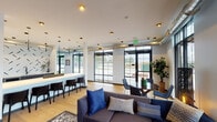 This single-floor space showcases a thoughtfully designed open-plan layout that seamlessly blends lounge, dining, and social areas. The interior is defined by a contemporary industrial-chic aesthetic, with exposed ductwork running along the ceiling contrasting against crisp white walls and warm light wood flooring throughout.

Floor-to-ceiling black-framed windows and glass doors dominate the exterior walls, flooding the space with abundant natural light and offering unobstructed outdoor views. The lounge area features a large sectional sofa in charcoal upholstery, accented with decorative pillows and anchored by a patterned area rug, while a small round dining table and modern chairs offer an intimate seating arrangement nearby.

A long, white-topped bar counter with dark cabinetry serves as a central focal point, accompanied by sleek pendant lighting and bar-height seating. Behind it, a bold graphic accent wall adds visual interest, complemented by a textured dark teal feature wall in the lounge area. A large arched mirror and potted fiddle-leaf fig tree further enrich the space. Structural columns are integrated naturally into the design, contributing to the loft-like, open ambiance of the overall space.