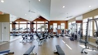 This fitness center is designed to offer a comprehensive workout experience in a bright, open environment. The space is anchored by dramatic vaulted ceilings with ceiling fans that promote airflow and comfort during exercise. Large windows line the walls, allowing generous amounts of natural light to fill the room and create an inviting, energizing atmosphere.

The facility is well-stocked with a diverse selection of cardio equipment, including stationary bikes, elliptical trainers, and treadmills, catering to a variety of aerobic fitness routines. Strength training is equally well represented, with a cable and pulley machine system, weight benches, and additional resistance equipment positioned throughout the space. The thoughtful layout ensures ample room to move between stations with ease.

The interior design features warm terracotta-toned accent walls that contrast with crisp white ceilings, lending the space a welcoming yet motivated ambiance. The flooring is a durable, cushioned surface suited for high-traffic fitness use. Informational posters are displayed on the walls, adding a functional and educational touch. Overall, this fitness center combines practical equipment variety with an appealing architectural design to support a well-rounded exercise experience.