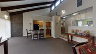 Located at 3674 Kent Rd in Stow, Ohio, this single-floor space makes a striking architectural impression with its soaring vaulted ceilings supported by bold, dark-stained exposed wood beams. Clerestory windows positioned high along the roofline allow natural light to pour into the open interior, enhancing the sense of height and volume throughout.

The main living area features a full-height brick accent wall that serves as a dramatic focal point, complementing the warm, neutral color palette and carpeted flooring found throughout the space. A comfortable seating arrangement with a leather sofa and coffee table anchors this area, with wall-mounted sconces providing ambient lighting.

A dedicated dining area accommodates a dark wood table with matching chairs, flanked by windows dressed with horizontal blinds and vertical shades that offer views to the exterior. The kitchen is accessed through an open passthrough and includes warm wood cabinetry, a black refrigerator, a microwave, and ample counter space, with a raised bar-height counter providing an additional casual seating option.

A stepped entryway leads to a secondary reception-style area featuring a console table and a pass-through window, suggesting a versatile, multi-use layout. The overall ambiance is warm and welcoming, combining rustic architectural elements with a functional, well-organized floor plan suited to a variety of uses.