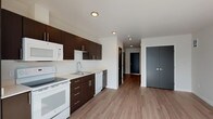 This contemporary single-floor unit offers a clean, modern aesthetic defined by a cohesive design palette and functional layout. The kitchen is a standout feature, outfitted with rich dark espresso cabinetry, sleek brushed metal hardware, and white countertops that provide a striking contrast. A full suite of white appliances includes a refrigerator, electric range, over-the-range microwave, dishwasher, and a stainless undermount sink — making it well-equipped for everyday use. Recessed lighting illuminates the space efficiently throughout.

Wood-look plank flooring extends seamlessly across the kitchen and into the open living area, creating a sense of continuity and warmth. The living space is bright and airy, benefiting from a large floor-to-ceiling window that invites ample natural light and provides views of the surrounding urban environment.

Charcoal-toned double doors and louvered interior doors add a modern, architectural touch to the unit's transitions between spaces. The bathroom maintains the same design language with a dark espresso vanity, white countertop, large mirror with overhead lighting, wood-look flooring, and a tub-shower combination with tiled surround. Overall, the unit presents a well-appointed, move-in ready space with a cohesive modern design throughout.