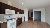 This contemporary single-floor unit offers a clean, modern aesthetic defined by a cohesive design palette and functional layout. The kitchen is a standout feature, outfitted with rich dark espresso cabinetry, sleek brushed metal hardware, and white countertops that provide a striking contrast. A full suite of white appliances includes a refrigerator, electric range, over-the-range microwave, dishwasher, and a stainless undermount sink — making it well-equipped for everyday use. Recessed lighting illuminates the space efficiently throughout.

Wood-look plank flooring extends seamlessly across the kitchen and into the open living area, creating a sense of continuity and warmth. The living space is bright and airy, benefiting from a large floor-to-ceiling window that invites ample natural light and provides views of the surrounding urban environment.

Charcoal-toned double doors and louvered interior doors add a modern, architectural touch to the unit's transitions between spaces. The bathroom maintains the same design language with a dark espresso vanity, white countertop, large mirror with overhead lighting, wood-look flooring, and a tub-shower combination with tiled surround. Overall, the unit presents a well-appointed, move-in ready space with a cohesive modern design throughout.