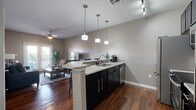 This single-floor residence offers a cohesive, open-concept layout that seamlessly connects the kitchen, dining, and living areas. Rich hardwood flooring runs throughout the main living spaces, complementing the neutral gray walls and creating a warm, inviting atmosphere.

The kitchen is a standout feature, equipped with dark espresso cabinetry, granite countertops, and a full suite of stainless steel appliances, including a refrigerator, range, and built-in microwave. A peninsula with bar seating accommodates three barstools and serves as both a prep space and casual dining area. Three pendant lights hang above, while track lighting provides additional illumination.

The living room receives natural light through French doors that open to an outdoor balcony. A ceiling fan overhead and rich hardwood flooring anchor the space comfortably.

The bedrooms feature plush carpeting, ceiling fans, and windows with plantation-style blinds. One bedroom also includes access to a private balcony through French doors.

The bathroom showcases dramatic marble-style tile on the floors and tub surround, paired with dark cabinetry, a granite vanity top, and a tub-shower combination, delivering a polished, contemporary finish.