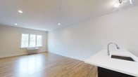 This single-floor space offers a clean, modern layout designed for comfortable everyday living. The open-concept floor plan connects the kitchen and living area in a seamless, flowing arrangement that maximizes usable space and promotes an airy atmosphere throughout.

The living area is finished with warm-toned hardwood flooring that runs continuously across the space, adding a sense of warmth and cohesion. Crisp white walls and a smooth white ceiling contribute to a bright, neutral aesthetic, while recessed lighting provides even, functional illumination throughout. A window along the far wall allows natural light to filter in, offering a view of lush greenery outdoors and further enhancing the open feel of the room. A wall-mounted HVAC unit ensures year-round climate control.

The kitchen area is defined by a sleek countertop overhang in white with a dark-toned base, creating a natural visual divide between the kitchen and living spaces. The contrast between the dark cabinetry and the light countertop adds a contemporary touch to the overall design.

The space is presented in a clean, move-in ready condition, with a minimalist finish palette that provides a versatile backdrop for a variety of interior styles and furnishing preferences.