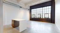 This single-floor loft space showcases a compelling blend of industrial heritage and contemporary design. The most striking architectural feature is the expansive factory-style window wall, framed in dark steel against painted brick, which floods the open living area with abundant natural light and offers views of the surrounding urban streetscape. Polished concrete floors run throughout, lending a sleek, cohesive look that complements the raw aesthetic.

Exposed ductwork and track lighting along the ceiling reinforce the industrial character, while crisp white walls provide a clean, gallery-like contrast. The open-plan layout creates a generous, flexible main area that flows naturally into the kitchen zone. The kitchen is outfitted with stainless steel appliances — including a range, dishwasher, microwave, and refrigerator — paired with rich wood-grain cabinetry, white countertops, and open shelving above. A central island provides additional workspace and casual seating.

A hallway lined with painted brick on one side leads to additional private rooms, including a bathroom with polished concrete floors and clean white finishes. The overall ambiance is contemporary yet grounded in industrial character, making the space well-suited for creative, live-work, or professional use.