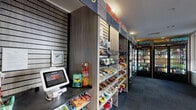 Located at 3009 M E Valentine Dr in Raleigh, NC, this single-floor Avenue C self-service market offers a modern and efficiently designed retail experience. The space is organized into two distinct zones: a well-stocked market area and an open adjoining lounge area. The market section features slatwall shelving units brimming with snack and convenience items, labeled by category — including "Recharge," "Nourish," "Chill," and "Hydrate" — alongside a row of glass-door refrigerators stocked with cold beverages, fresh food, and drinks. A self-checkout kiosk with a "Simply Scan" tablet interface allows for a seamless, cashierless shopping experience. The adjoining open area is finished with commercial-grade carpet tiles in a muted blue-grey tone and warm yellow accent walls adorned with a trio of digital-art canvas prints. Large corner windows flood this area with natural light and offer views of the surrounding urban streetscape. Recessed ceiling lighting throughout maintains a clean, functional ambiance. An inspirational wall-mounted artwork reading "inspire, use imagination, reinvent" adds a motivational touch to the space. The overall layout is intuitive and accessible, designed to serve the needs of a busy community efficiently.