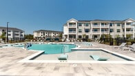 Located at 420 Angler Dr in Wilmington, NC, this two-floor residential community delivers a resort-style living experience with standout outdoor amenities and coastal-inspired architecture. The exterior presents a classic coastal design, featuring white lap siding, dark-trimmed windows, pitched rooflines, and covered balconies that lend a refined, cohesive aesthetic throughout the multi-building complex.

At the heart of the community lies a generously sized freeform swimming pool with a sun shelf and in-water loungers, ideal for relaxation in the warm North Carolina climate. A separate wading or spa area complements the main pool, while a spacious tiled pool deck is lined with lounge chairs, offering ample room for sunbathing and outdoor entertaining. Under a distinctive round-roofed covered pavilion with a standing-seam metal roof, residents can enjoy shaded outdoor seating, creating an inviting gathering space. Additional outdoor amenities include cabana-style seating areas, an outdoor grill station with dining furniture, and meticulously maintained landscaping with mature shrubbery and greenery that soften the hardscape. The community is enclosed by a black iron perimeter fence, providing both security and a polished finish to the overall grounds.