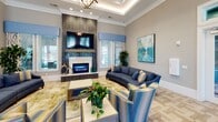 This sophisticated single-floor community or leasing facility is designed with upscale finishes and thoughtful details throughout. The grand reception area makes an immediate impression with its coffered tray ceiling adorned with exposed wood beams, a striking multi-arm chandelier, and a curved front desk finished in deep blue tones. The soaring entryway features large-format stone tile flooring, floor-to-ceiling glass panels that flood the space with natural light, and curated wall art flanked by lush potted plants. A comfortable lounge area offers a welcoming retreat, complete with a floor-to-ceiling herringbone tile fireplace surround, a wall-mounted television, a plush blue sofa, and patterned accent chairs set atop a chevron-patterned hardwood floor. A dedicated business center/lounge is furnished with a built-in workstation set against a dramatic marble accent wall, providing a quiet space for focused work. A secondary lounge area, accessible through glass-paned double doors, seamlessly connects the space. The kitchenette features dark cabinetry, a vibrant mosaic tile backsplash, stainless steel appliances, and a communal high-top table bathed in natural light. Throughout, tray ceilings with crown molding, abundant recessed lighting, and cohesive coastal-inspired décor create a polished and inviting ambiance.