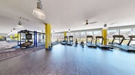 Located at 1 Merrimack Plaza in Lowell, Massachusetts, this expansive fitness center offers a generously proportioned, single-floor exercise space designed to accommodate a wide range of workout needs. The interior makes a strong visual statement with bold yellow columns and accent walls that punctuate the bright, white ceiling, creating an energetic and motivating atmosphere throughout.

The cardio zone features a row of treadmills, ellipticals, stationary bikes, and a rowing machine, all positioned along a continuous bank of floor-to-ceiling windows that frame views of the surrounding Lowell streetscape, flooding the space with natural light. Ceiling fans and industrial-style pendant lighting ensure year-round comfort and visibility.

The strength and functional training area is equally well-appointed, with cable machines, weight benches, free weights, plyometric boxes, and a multi-station functional training rig. A textured stone accent wall adds architectural character to this zone, while large mirrors along the walls enhance both the sense of space and workout functionality.

Flooring transitions between durable rubber in the cardio areas and a stylish herringbone-patterned carpet tile in the strength zones. Multiple double-door entry points provide convenient access. The overall design is modern, clean, and purpose-built to support a comprehensive fitness experience.