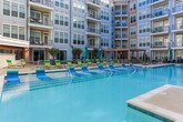 Located in the prestigious 30305 zip code of Atlanta, Georgia, this outdoor amenity space is centered around a generously sized swimming pool set within a striking multi-story courtyard. The pool's crystal-clear, light-blue water is complemented by in-pool lounge chairs in vibrant teal and aqua tones, inviting residents to relax directly in the water. The surrounding deck is finished in a neutral concrete surface and furnished with an array of green and teal sling-style chaise lounges, dining tables, and chairs, all arranged to encourage both sunbathing and social gatherings. Teal market umbrellas provide shaded retreats throughout the deck area. Lush potted greenery and manicured plantings are thoughtfully placed to add natural texture and color to the space. The courtyard is enclosed on multiple sides by the building's multi-story facade, which features large windows, private balconies, and a mix of brick and contemporary paneling — creating a sense of privacy and enclosure while maintaining an open-air feel. String lights are visible along the building's exterior, adding a warm ambiance for evening enjoyment. This well-designed outdoor space offers a resort-like retreat in the heart of Atlanta.