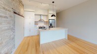 Located at 320 S 6th St in Milwaukee's Walker's Point neighborhood, this single-floor unit blends industrial character with modern finishes throughout its thoughtfully designed layout. The open-concept living and kitchen area serves as the heart of the space, where light hardwood flooring stretches across a generous footprint. A striking exposed cream brick wall adds historic texture and visual warmth, contrasting beautifully with the clean, white contemporary finishes found throughout.

The kitchen is well-appointed with white cabinetry, a large central island featuring a stainless steel sink and white countertops, stainless steel appliances including a range with a wall-mounted hood vent, and a French door refrigerator. Rustic wood floating shelves and a subway tile backsplash add character to the culinary space, while a pendant light anchors the island.

Additional rooms feature plush patterned carpeting, exposed cream brick accent walls, and neutral painted walls, creating versatile spaces suitable for sleeping or office use. Large floor-to-ceiling windows and a glass door in one room invite substantial natural light and provide views of the surrounding Milwaukee cityscape.

The bathroom is finished with light linear tile flooring, a wall-mounted grab bar, a clean white toilet, and warm wood-toned cabinetry, delivering a modern and functional design.