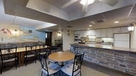 This single-floor space is thoughtfully arranged to accommodate group gatherings, combining a comfortable dining area with a fully equipped kitchen. The dining section features multiple tables, including a round table with ornate wrought-iron chairs and a longer rectangular table with additional seating, making it well-suited for communal meals or events. The flooring is carpeted throughout the dining area, adding a degree of warmth to the functional setting.

The kitchen is separated from the dining area by a stone-faced breakfast bar, which serves as both a visual divider and a practical surface. The kitchen is outfitted with white cabinetry, a microwave, a sink, ample counter space, and additional appliances, providing everything needed for meal preparation and service.

The drop ceiling features recessed lighting and a ceiling fan, ensuring adequate illumination across the space. A decorative mural depicting a scenic landscape adds a touch of character to one wall, while an exit-lit doorway provides access to an adjoining corridor or room. The overall layout is practical and welcoming, designed to support a variety of communal activities in an organized and accessible single-level environment.
