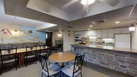 This single-floor space is thoughtfully arranged to accommodate group gatherings, combining a comfortable dining area with a fully equipped kitchen. The dining section features multiple tables, including a round table with ornate wrought-iron chairs and a longer rectangular table with additional seating, making it well-suited for communal meals or events. The flooring is carpeted throughout the dining area, adding a degree of warmth to the functional setting.

The kitchen is separated from the dining area by a stone-faced breakfast bar, which serves as both a visual divider and a practical surface. The kitchen is outfitted with white cabinetry, a microwave, a sink, ample counter space, and additional appliances, providing everything needed for meal preparation and service.

The drop ceiling features recessed lighting and a ceiling fan, ensuring adequate illumination across the space. A decorative mural depicting a scenic landscape adds a touch of character to one wall, while an exit-lit doorway provides access to an adjoining corridor or room. The overall layout is practical and welcoming, designed to support a variety of communal activities in an organized and accessible single-level environment.