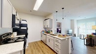 Located at 221 Waldron St in West Lafayette, Indiana, this one-bedroom, one-bathroom unit offers a practical and modern layout across a single floor. The open-concept kitchen and living area serves as the heart of the space, featuring light wood-style flooring, crisp white walls, and a seamless flow between cooking and relaxing zones. The kitchen is well-appointed with gray shaker-style cabinetry, light granite countertops, a subway tile backsplash, stainless steel appliances including a range, microwave, and refrigerator, and a peninsula island with pendant lighting and bar seating — ideal for casual dining or studying. A skylight overhead adds a bright, airy feel to the kitchen area. The living room benefits from a large sliding glass door with vertical blinds, allowing ample natural light and outdoor views of the surrounding greenery. The carpeted bedroom is a comfortable retreat, accommodating a full bed with under-bed storage and a dedicated desk area for studying or working. Two closets provide generous storage throughout the unit. The bathroom includes a full tub-shower combo with clean white finishes. Light wood-style flooring runs throughout the main living areas, tying the space together with a cohesive, contemporary aesthetic.