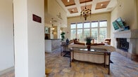 This single-floor space in Castle Rock, Colorado, presents a refined and elegantly appointed interior defined by dramatic architectural details. The great room commands immediate attention with its soaring ceiling height, coffered tray ceiling accents, and a striking wrought-iron chandelier that anchors the space. A bank of large, multi-pane windows along one wall floods the interior with natural light while offering views of the surrounding greenery.

Arched doorways supported by substantial columns lend a Mediterranean-inspired character throughout. The flooring is laid in warm, earth-toned stone tiles that complement the neutral cream wall tones. A prominent fireplace with a crisp white surround and decorative wrought-iron screen serves as a focal centerpiece, accompanied by a wall-mounted television above.

A dedicated wet bar area is tucked to one side, equipped with dark countertops, white cabinetry with glass-front upper doors, a stone tile backsplash, a sink, and a built-in beverage refrigerator — all accented by ornate wall sconces. The reception or entry area features a tiled counter and pendant lighting. Throughout the space, decorative ironwork details, tasteful framed artwork, and layered lighting contribute to a warm, upscale ambiance suited for both social gatherings and everyday use.