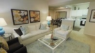 This single-floor residence showcases an open-concept design that effortlessly connects the living, dining, and kitchen areas into one cohesive and inviting space. The living area serves as the centerpiece, featuring plush white upholstered seating — including a sofa and armchair — arranged around a sleek glass-top coffee table. Soft aqua and floral accent pillows add a refreshing pop of color against the neutral palette, complementing the light-toned area rug underfoot. Two large floral canvas artworks adorn the wall, lending warmth and character to the space. A blue ceramic table lamp adds a stylish accent and soft ambient lighting. Just beyond the living area, the dining space accommodates a full table and chair set, creating a natural flow for entertaining. The kitchen is partially visible with light cabinetry and a counter that transitions smoothly from the dining area. Throughout the space, recessed lighting and warm-toned walls contribute to a clean, welcoming ambiance. The open layout maximizes the sense of space, making the interior feel airy and well-proportioned. The overall design aesthetic is contemporary coastal, blending comfort with understated elegance.