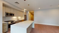 Located at 3193 Washington St in Boston's Jamaica Plain neighborhood, this thoughtfully designed single-floor residence combines modern aesthetics with functional living. The open-concept layout connects the kitchen and living area seamlessly, creating a spacious and airy atmosphere bathed in natural light from a bank of large windows.

The kitchen is a standout feature, showcasing sleek flat-front cabinetry in a soft greige tone, a full suite of stainless steel appliances including a gas range, built-in microwave, dishwasher, and a full-size refrigerator. A geometric tile backsplash and crisp white quartz countertops add refined detail, while pendant lighting over the generous kitchen island provides both task lighting and visual interest.

Warm hardwood flooring flows consistently throughout the main living spaces and bedrooms, lending cohesion and warmth to the overall design. The bedrooms are well-proportioned, with one featuring a large closet with sliding doors and a high transom window for natural light. An additional room offers flexibility for use as a second bedroom or home office.

The bathroom is clean and efficiently designed, featuring a full soaking tub with shower, neutral tile flooring, and a white vanity. Recessed lighting, clean white walls, and louvered interior doors complete the modern, polished character of this well-appointed Boston residence.