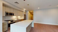 Located at 3193 Washington St in Boston's Jamaica Plain neighborhood, this thoughtfully designed single-floor residence combines modern aesthetics with functional living. The open-concept layout connects the kitchen and living area seamlessly, creating a spacious and airy atmosphere bathed in natural light from a bank of large windows.

The kitchen is a standout feature, showcasing sleek flat-front cabinetry in a soft greige tone, a full suite of stainless steel appliances including a gas range, built-in microwave, dishwasher, and a full-size refrigerator. A geometric tile backsplash and crisp white quartz countertops add refined detail, while pendant lighting over the generous kitchen island provides both task lighting and visual interest.

Warm hardwood flooring flows consistently throughout the main living spaces and bedrooms, lending cohesion and warmth to the overall design. The bedrooms are well-proportioned, with one featuring a large closet with sliding doors and a high transom window for natural light. An additional room offers flexibility for use as a second bedroom or home office.

The bathroom is clean and efficiently designed, featuring a full soaking tub with shower, neutral tile flooring, and a white vanity. Recessed lighting, clean white walls, and louvered interior doors complete the modern, polished character of this well-appointed Boston residence.