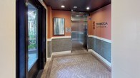 The Tribeca Urban lobby presents a well-appointed, single-floor entry experience that balances contemporary design with practical resident amenities. The entrance corridor is immediately distinguished by its herringbone-patterned brick flooring, which leads residents and guests through a thoughtfully organized space. Warm terracotta-orange upper walls contrast with textured gray wainscoting and crisp white baseboards, creating a layered, inviting color palette throughout.

A stainless steel elevator serves as a central architectural focal point, positioned prominently within the lobby and flanked by curated abstract artwork that adds a gallery-like quality to the surroundings. Recessed ceiling lighting throughout ensures consistent, well-distributed illumination across all areas.

Adjacent to the main lobby, a dedicated mailroom houses an extensive bank of individually numbered resident mailboxes finished in dark bronze, neatly organized in a corner alcove with coordinating herringbone flooring. A separate package area features a generous granite-topped counter with dark cabinetry below, offering a refined and functional space for parcel management.

A large glass window panel provides a view into an adjoining corridor lined with light-colored tile, further enhancing the open, connected feel of the space. Lush succulent arrangements and framed landscape art contribute to the lobby's polished, welcoming ambiance. A green building certification plaque is also displayed, reflecting a commitment to sustainability.