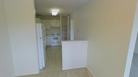 This single-floor residence offers a practical and straightforward layout designed for comfortable everyday living. The kitchen is a standout functional space, outfitted with white appliances including a full-size refrigerator and a freestanding electric range with an overhead ventilation hood. White upper cabinetry provides ample storage, while light-toned floor tiles lend a clean, bright aesthetic to the cooking area. A built-in open shelving unit adds additional storage and organizational flexibility alongside the cabinetry. A white peninsula wall separates the kitchen from the adjacent living space, creating a natural transition between the two areas while maintaining an open, connected feel. The living area features light-colored flooring that complements the overall neutral palette of the home. The interiors are finished in soft, warm tones throughout, contributing to a cohesive and understated ambiance. Flush ceiling lighting provides consistent illumination across the space. The overall design emphasizes utility and simplicity, making it a well-suited space for those seeking a clean, move-in-ready environment with all essential features thoughtfully incorporated into a compact, efficient footprint.