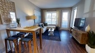 This single-floor residence showcases a thoughtfully designed open-concept layout that seamlessly connects the dining and living areas. Rich, dark hardwood-style flooring runs throughout, lending warmth and continuity to the space. Neutral gray walls provide a versatile backdrop that complements a range of décor styles.

The living area is anchored by large windows dressed with sheer curtains and horizontal blinds, allowing generous natural light to fill the room. A glass-paneled door opens to what appears to be a private balcony, extending the living space outdoors and offering views of the surrounding greenery. The room comfortably accommodates a sectional sofa arrangement centered around a modern glass coffee table atop a deep navy area rug.

Adjacent to the living area, a raised dining table with bar-height seating defines a casual dining space, making it ideal for everyday meals or entertaining. A floor lamp and wall art contribute to the polished yet relaxed ambiance of the space. Overall, the layout feels open and functional, with a clean, contemporary aesthetic throughout.