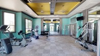 The fitness center offers a bright and energizing workout environment defined by its bold design choices and functional layout. The space is finished with vivid teal walls that create an invigorating atmosphere, complemented by a distinctive tray ceiling accented in yellow with pendant lighting that adds visual interest and warmth overhead. Recessed lighting throughout ensures the space is well-lit at all times.

The floor is covered in durable, patterned carpet tiles suited for high-traffic use. A large mirror spans a central wall, visually expanding the space and aiding exercisers in monitoring their form. Natural light enters through windows along one wall, keeping the environment open and welcoming.

Equipment includes cardio machines such as stationary bikes and an elliptical, as well as a dumbbell rack with a weight bench for strength training. Additional equipment is visible in the mirrored section, suggesting a functional layout designed to accommodate a variety of workout routines. A wall-mounted television is also present. French-style glass doors provide access to an adjacent area, further enhancing the open feel of the space.