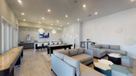 This contemporary single-floor amenity space is thoughtfully designed to serve as a social hub, blending stylish aesthetics with functional entertainment areas. Wood-look tile flooring flows throughout, unifying the open layout beneath recessed lighting and statement chandelier fixtures that add a touch of elegance.

The lounge area anchors one end of the space, featuring plush gray sectional sofas and geometric accent tables arranged around a large wall-mounted flat-screen TV set against a bold dark wood accent wall. Circular decorative wall elements add an artistic touch to the adjacent seating areas, where additional modular lounge furniture invites casual conversation.

At the center of the room, a pool table serves as a focal point for recreation, complemented by a shuffleboard table nearby. High-top bistro tables with bar-height seating provide intimate spots for smaller gatherings throughout the space.

The wet bar and kitchen area is a standout feature, showcasing a dramatic veined marble island with bar seating, dark cabinetry, a patterned geometric tile backsplash, an undermount sink, and a dedicated Starbucks-branded coffee and tea station. A wall-mounted TV above the bar adds to the entertaining atmosphere. Glass-paneled doors along one wall invite natural light and connect the interior to the outdoors.