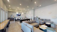 This contemporary single-floor amenity space is thoughtfully designed to serve as a social hub, blending stylish aesthetics with functional entertainment areas. Wood-look tile flooring flows throughout, unifying the open layout beneath recessed lighting and statement chandelier fixtures that add a touch of elegance.

The lounge area anchors one end of the space, featuring plush gray sectional sofas and geometric accent tables arranged around a large wall-mounted flat-screen TV set against a bold dark wood accent wall. Circular decorative wall elements add an artistic touch to the adjacent seating areas, where additional modular lounge furniture invites casual conversation.

At the center of the room, a pool table serves as a focal point for recreation, complemented by a shuffleboard table nearby. High-top bistro tables with bar-height seating provide intimate spots for smaller gatherings throughout the space.

The wet bar and kitchen area is a standout feature, showcasing a dramatic veined marble island with bar seating, dark cabinetry, a patterned geometric tile backsplash, an undermount sink, and a dedicated Starbucks-branded coffee and tea station. A wall-mounted TV above the bar adds to the entertaining atmosphere. Glass-paneled doors along one wall invite natural light and connect the interior to the outdoors.