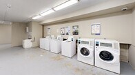 Located at 301-341 Traynor Ave in Kitchener, Ontario, this shared laundry facility offers a practical and well-equipped space designed to serve a residential community. The facility is laid out across a single floor and comprises a main laundry room along with an adjoining utility/entry area.

The main laundry room is generously sized, accommodating a substantial row of top-loading washing machines and front-loading washers along one wall, paired with a matching lineup of front-loading dryers along the opposite wall. The machines are modern and coin- or card-operated, as indicated by informational signage and posted instructions displayed throughout the room. Overhead fluorescent lighting illuminates the entire space efficiently. The floor is finished in a durable concrete-style surface suited for high-traffic use.

The adjoining entry and utility area features the same practical flooring and neutral cream-toned walls. A stainless steel utility table provides a convenient surface for sorting or folding laundry, while a notice board with posted information is mounted on the wall. This area also connects to additional utility infrastructure, including what appears to be a utility sink.

Overall, the facility presents a clean, functional, and straightforward environment designed with convenience and practicality in mind, providing residents with all essential amenities for on-site laundry needs.